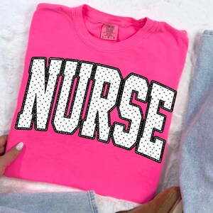 Peut inclure: Un t-shirt rose vif avec le mot "NURSE" en grandes lettres blanches &agrave; pois noirs et un contour noir. Le t-shirt est pli&eacute;, et une partie d'un jean bleu est visible.