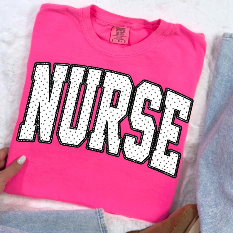 Peut inclure: Un t-shirt rose vif avec le mot "NURSE" en grandes lettres blanches &agrave; pois noirs et un contour noir. Le t-shirt est pli&eacute;, et une partie d'un jean bleu est visible.