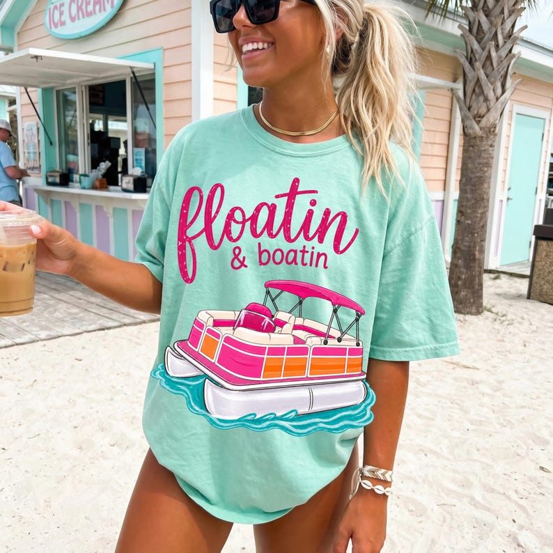 May include: A light green t-shirt with the words "floatin & boatin" in pink script above a pink and orange pontoon boat graphic. The shirt is worn on a beach.