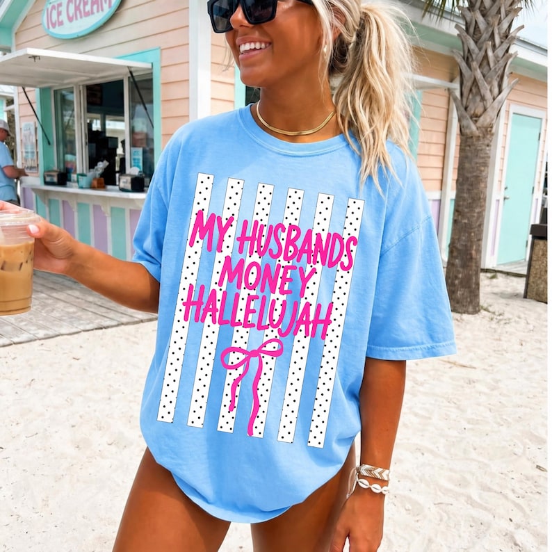 May include: A light blue t-shirt featuring a graphic print of vertical white stripes with black dots. The text "MY HUSBANDS MONEY HALLELUJAH" is printed in bright pink letters across the center, accompanied by a pink line-drawn bow graphic.