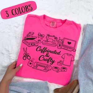 Op de afbeelding: Een felroze t-shirt met de tekst "Caffeinated & Crafty" en zwarte illustraties van knutselgereedschap, waaronder een naaimachine, laptop en schaar. Het ontwerp bevat ook de tekst "3 COLORS" in een roze ovaal.