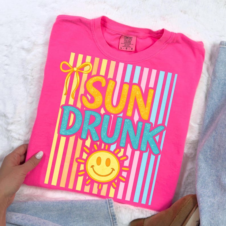 May include: A bright pink t-shirt with the words "SUN DRUNK" in yellow and teal, with a smiling sun graphic. The shirt has a striped design and a yellow bow. The shirt is folded on a white surface.