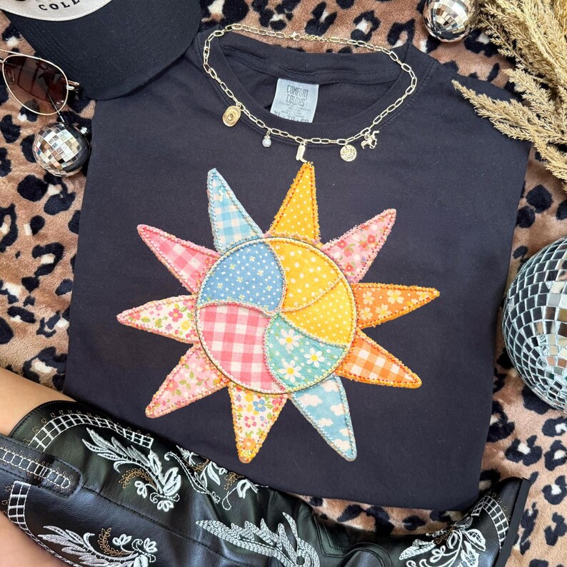 Puede incluir: Sudadera negra de cuello redondo con un dise&ntilde;o de sol de patchwork colorido. El patr&oacute;n de sol incluye varias telas pastel. Un collar de cadena dorada con dijes descansa sobre la camisa. Gafas de sol y gorra negra en el fondo.