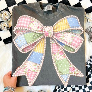 May include: Dark gray sweatshirt featuring a large, colorful bow design. The bow is made of patchwork fabric in pastel colors, including pink, yellow, blue, and green, with a pearl-like trim. The sweatshirt has a crew neck and long sleeves.