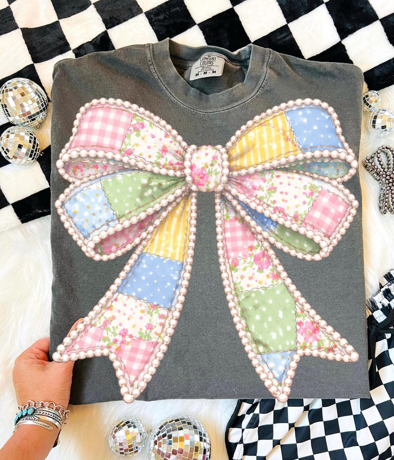 May include: Dark gray sweatshirt featuring a large, colorful bow design. The bow is made of patchwork fabric in pastel colors, including pink, yellow, blue, and green, with a pearl-like trim. The sweatshirt has a crew neck and long sleeves.