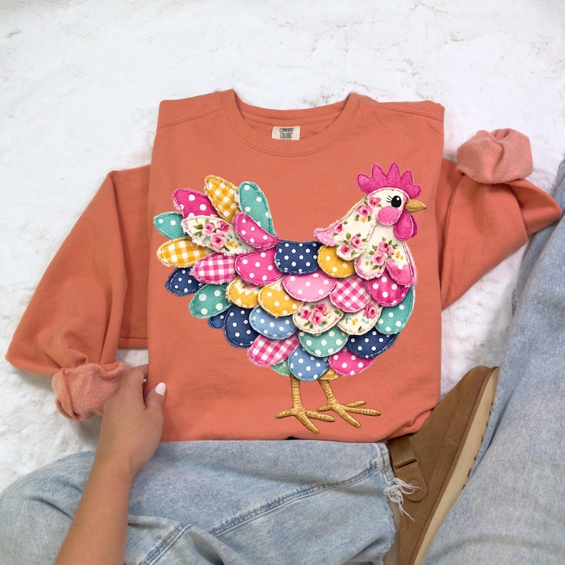 May include: An orange crew neck sweatshirt featuring a colorful, patchwork-style chicken design. The chicken has a floral patterned head and a tail with polka dots, gingham, and floral patterns. The sweatshirt has long sleeves and a relaxed fit.