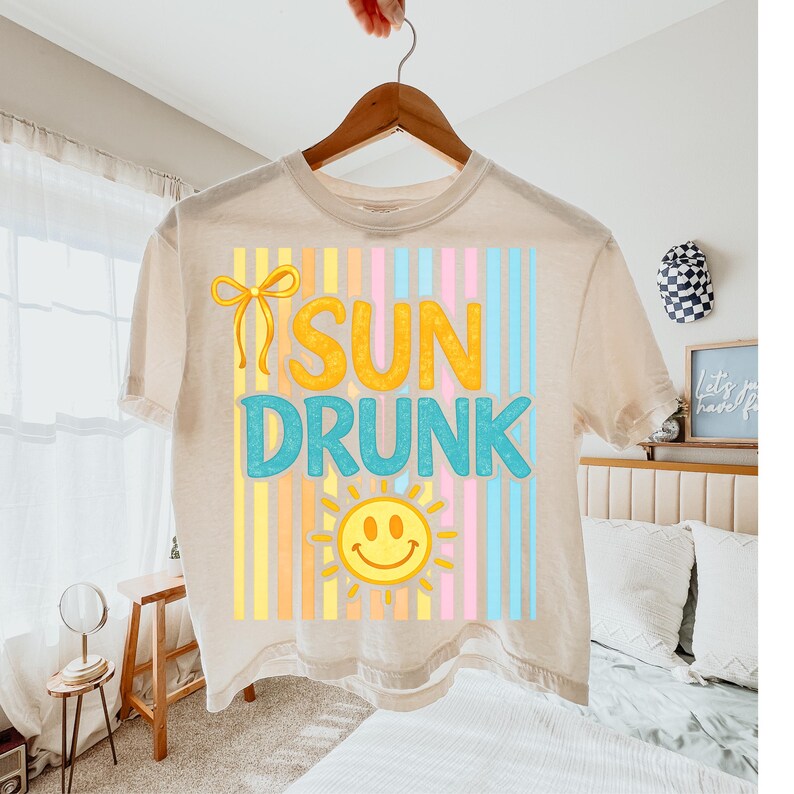 May include: Beige t-shirt with the words "SUN DRUNK" in yellow and blue, with a smiling sun graphic. The design features vertical stripes in pastel colors. The shirt is hanging on a wooden hanger.