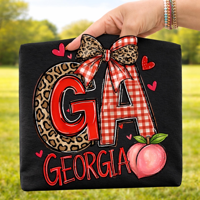Puede incluir: Bolso tote negro con las letras "GA" en rojo y estampado de leopardo, un lazo de cuadros y un gr&aacute;fico de melocot&oacute;n. La palabra "GEORGIA" est&aacute; escrita en rojo debajo. Peque&ntilde;os corazones rojos est&aacute;n dispersos alrededor del dise&ntilde;o.