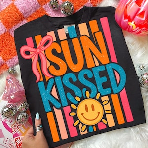May include: Black crew neck sweatshirt with the words "SUN KISSED" in orange and blue, a smiling sun graphic, and a pink bow. The sweatshirt has vertical stripes in orange, pink, and peach.
