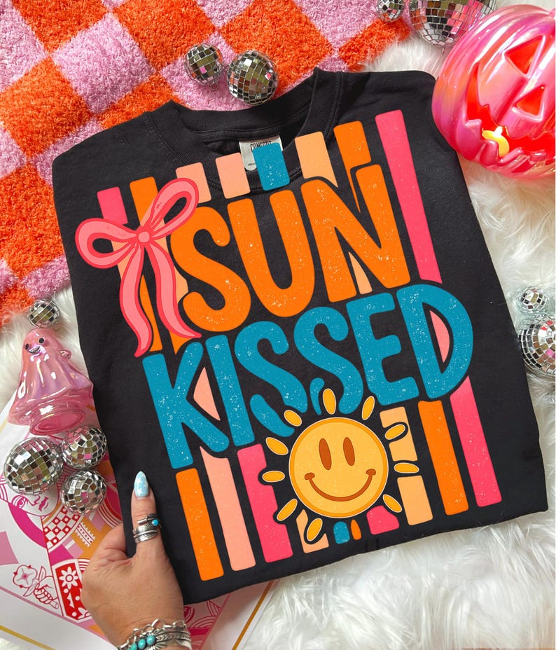 May include: Black crew neck sweatshirt with the words "SUN KISSED" in orange and blue, a smiling sun graphic, and a pink bow. The sweatshirt has vertical stripes in orange, pink, and peach.