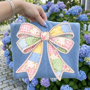 May include: A light blue clutch featuring a large, decorative bow. The bow is made of patchwork fabric in pastel colors, including pink, yellow, green, and blue, with a pearl-like trim. The background shows blue and purple hydrangeas.
