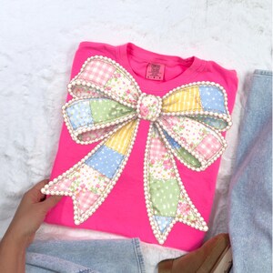 May include: A bright pink t-shirt featuring a large, decorative bow. The bow is made of patchwork fabric in pastel colors, including pink floral, blue polka dots, and yellow gingham. The bow is outlined with white pearls.