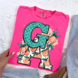 May include: A vibrant pink sweatshirt with a large teal letter "G" in a leopard print. The letter "A" is filled with peach illustrations. Peach blossoms and peaches surround the letters, creating a summery design.