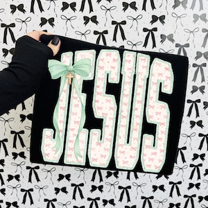 May include: Black clutch with the word "JESUS" in large, white, bow-patterned letters. A light green bow and gold cross are attached to the side. The background is a pattern of black and white bows.