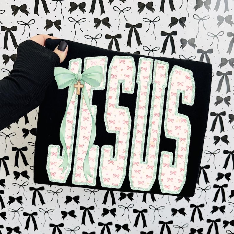 May include: Black clutch with the word "JESUS" in large, white, bow-patterned letters. A light green bow and gold cross are attached to the side. The background is a pattern of black and white bows.