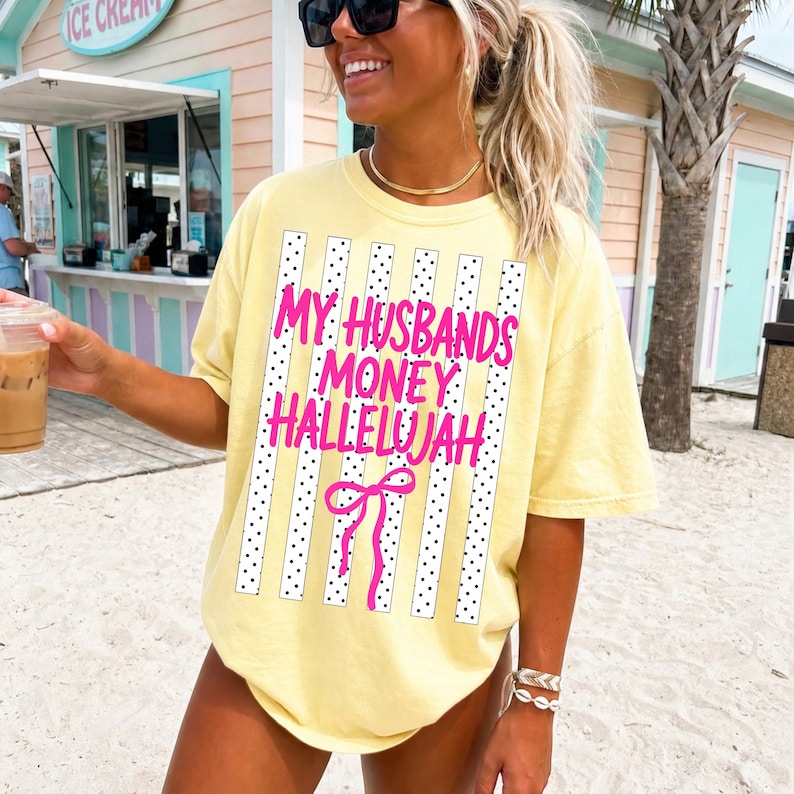 May include: A pale yellow short-sleeve t-shirt featuring a graphic print of vertical white stripes with black dots. The text reads MY HUSBANDS MONEY HALLELUJAH in bright pink letters, accompanied by a pink line-drawn bow graphic centered below.