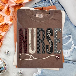 May include: A brown short-sleeve t-shirt featuring the word NURSE in large block letters, each with a different pattern: leopard print, spider webs, black and gray checkers, skeletons, and white polka dots, with a stethoscope graphic below.