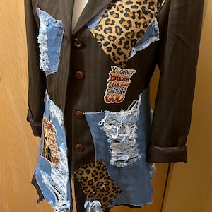 May include: A brown pinstripe blazer with denim patches and leopard print accents. The blazer features distressed denim patches, leopard print fabric, and a patch with the text "You cannot break a person who gets their strength from God."