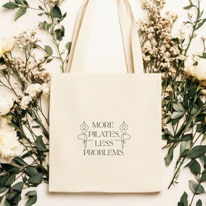 Eine beigefarbene Stofftasche liegt flach auf einem Hintergrund aus weißen Rosen, zarten Blüten und grünen Pflanzen. In der Mitte der Tasche steht der Spruch „More Pilates, Less Problems.“ in eleganter, minimalistischer Typografie, ergänzt durch zwei filigrane Pilates-Silhouetten. Die ästhetische, natürliche Szene vermittelt Ruhe, Wellness und einen aktiven, gesunden Lifestyle – ideal für Pilates-Liebhaber und nachhaltige Geschenkideen.