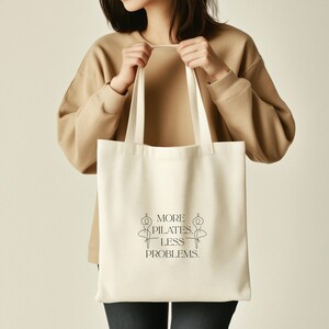 Eine beigefarbene Stofftasche liegt flach auf einem Hintergrund aus weißen Rosen, zarten Blüten und grünen Pflanzen. In der Mitte der Tasche steht der Spruch „More Pilates, Less Problems.“ in eleganter, minimalistischer Typografie, ergänzt durch zwei filigrane Pilates-Silhouetten. Die ästhetische, natürliche Szene vermittelt Ruhe, Wellness und einen aktiven, gesunden Lifestyle – ideal für Pilates-Liebhaber und nachhaltige Geschenkideen.