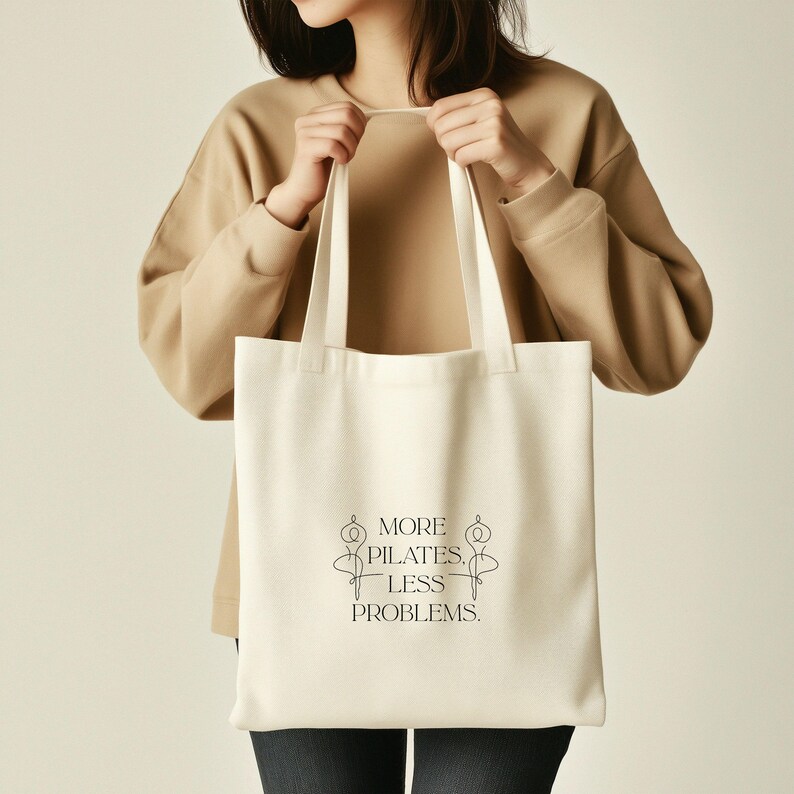 Eine beigefarbene Stofftasche liegt flach auf einem Hintergrund aus weißen Rosen, zarten Blüten und grünen Pflanzen. In der Mitte der Tasche steht der Spruch „More Pilates, Less Problems.“ in eleganter, minimalistischer Typografie, ergänzt durch zwei filigrane Pilates-Silhouetten. Die ästhetische, natürliche Szene vermittelt Ruhe, Wellness und einen aktiven, gesunden Lifestyle – ideal für Pilates-Liebhaber und nachhaltige Geschenkideen.