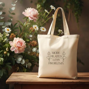 Eine beigefarbene Stofftasche liegt flach auf einem Hintergrund aus weißen Rosen, zarten Blüten und grünen Pflanzen. In der Mitte der Tasche steht der Spruch „More Pilates, Less Problems.“ in eleganter, minimalistischer Typografie, ergänzt durch zwei filigrane Pilates-Silhouetten. Die ästhetische, natürliche Szene vermittelt Ruhe, Wellness und einen aktiven, gesunden Lifestyle – ideal für Pilates-Liebhaber und nachhaltige Geschenkideen.