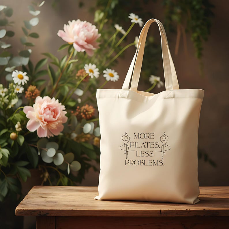 Eine beigefarbene Stofftasche liegt flach auf einem Hintergrund aus weißen Rosen, zarten Blüten und grünen Pflanzen. In der Mitte der Tasche steht der Spruch „More Pilates, Less Problems.“ in eleganter, minimalistischer Typografie, ergänzt durch zwei filigrane Pilates-Silhouetten. Die ästhetische, natürliche Szene vermittelt Ruhe, Wellness und einen aktiven, gesunden Lifestyle – ideal für Pilates-Liebhaber und nachhaltige Geschenkideen.