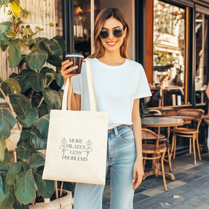 Eine beigefarbene Stofftasche liegt flach auf einem Hintergrund aus weißen Rosen, zarten Blüten und grünen Pflanzen. In der Mitte der Tasche steht der Spruch „More Pilates, Less Problems.“ in eleganter, minimalistischer Typografie, ergänzt durch zwei filigrane Pilates-Silhouetten. Die ästhetische, natürliche Szene vermittelt Ruhe, Wellness und einen aktiven, gesunden Lifestyle – ideal für Pilates-Liebhaber und nachhaltige Geschenkideen.