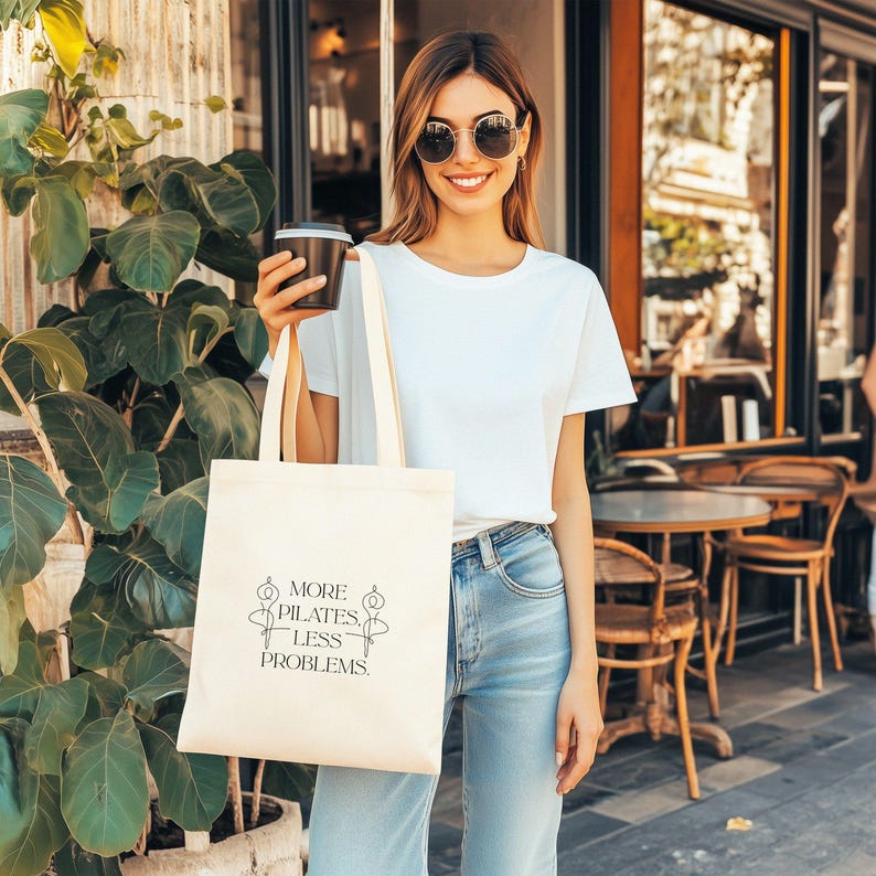 Eine beigefarbene Stofftasche liegt flach auf einem Hintergrund aus weißen Rosen, zarten Blüten und grünen Pflanzen. In der Mitte der Tasche steht der Spruch „More Pilates, Less Problems.“ in eleganter, minimalistischer Typografie, ergänzt durch zwei filigrane Pilates-Silhouetten. Die ästhetische, natürliche Szene vermittelt Ruhe, Wellness und einen aktiven, gesunden Lifestyle – ideal für Pilates-Liebhaber und nachhaltige Geschenkideen.