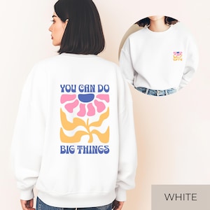 Könnte beinhalten: Weißes Sweatshirt mit einem Blumendesign auf dem Rücken, das eine rosa Blume, einen blauen Mittelpunkt und gelbe Akzente aufweist. Der Text "YOU CAN DO BIG THINGS" befindet sich über und unter der Blume. Eine kleinere Version des Designs befindet sich auf der Vorderseite.