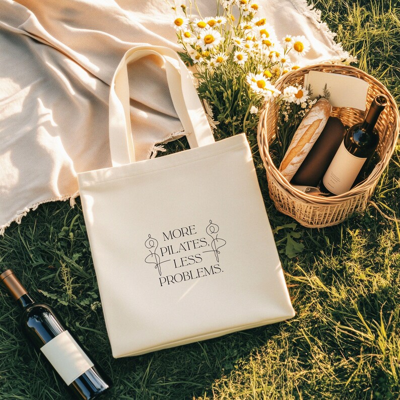 Eine beigefarbene Stofftasche liegt flach auf einem Hintergrund aus weißen Rosen, zarten Blüten und grünen Pflanzen. In der Mitte der Tasche steht der Spruch „More Pilates, Less Problems.“ in eleganter, minimalistischer Typografie, ergänzt durch zwei filigrane Pilates-Silhouetten. Die ästhetische, natürliche Szene vermittelt Ruhe, Wellness und einen aktiven, gesunden Lifestyle – ideal für Pilates-Liebhaber und nachhaltige Geschenkideen.