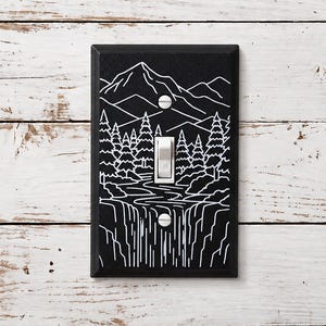May include: A rectangular black light switch cover plate featuring a white line art illustration of a mountain range, a dense forest of pine trees, and a flowing waterfall. The design includes a central white toggle switch and two silver screws.
