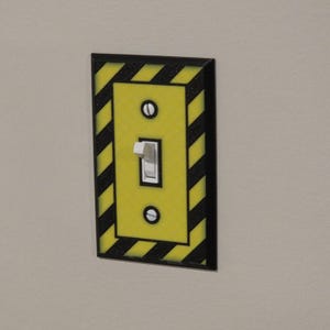 May include: A rectangular light switch cover plate featuring a yellow center panel and a black and yellow diagonal hazard stripe border. The plate is secured with two flathead screws and contains a single white toggle switch in the center.