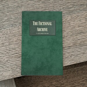 Könnte beinhalten: Ein grünes Buch mit dem Titel "The Fictional Archive: YA Readers Edition" liegt auf einer Holzoberfläche. Das Buch hat einen dunkelgrünen Einband mit einem rechteckigen Etikett in der Mitte, das den Titel in weißem Text anzeigt.
