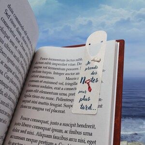 May include: A white bookmark with the text "Je prends des notes pour plus tard..." and a red tie illustration, inserted into an open book. The book has brown covers and pages with printed text.