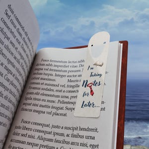 Op de afbeelding: Een bladwijzer met de tekst "I'm Taking Notes for Later..." is in een boek geplaatst. De bladwijzer heeft een kleine illustratie van een boek en een rode stropdas. Het boek heeft bruine kaften en witte pagina's.