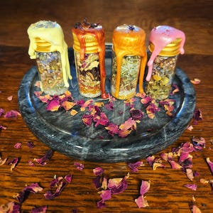 May include: Four small glass bottles filled with dried herbs and flowers, each topped with a different color of dripped wax: yellow, orange-red, orange, and pink. The bottles sit on a dark gray, round stone tray, surrounded by scattered dried rose petals.