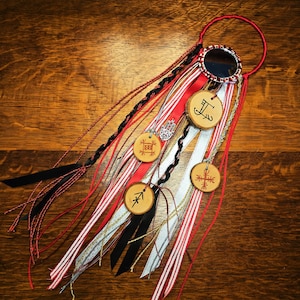 May include: A handcrafted hair accessory featuring a circular mirror and a red loop. It has multiple ribbons in red, white, black, and silver, along with wooden charms with etched symbols and a silver charm. The accessory is displayed on a wooden surface.