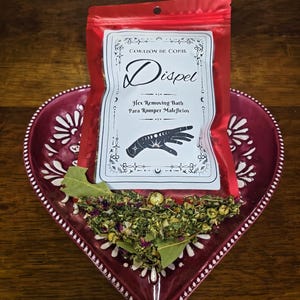 May include: A red bag labeled "Dispel" with text in Spanish, resting on a heart-shaped, burgundy dish. The dish holds dried herbs and bay leaves. The bag's label features a hand with crescent moons.