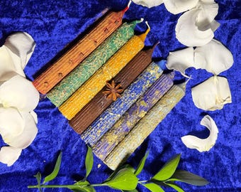 May include: An assortment of colorful, decorative candles arranged on a deep blue velvet surface. The candles are various colors, including orange, green, yellow, brown, blue, and gold. White flower petals and green leaves are scattered around the candles.