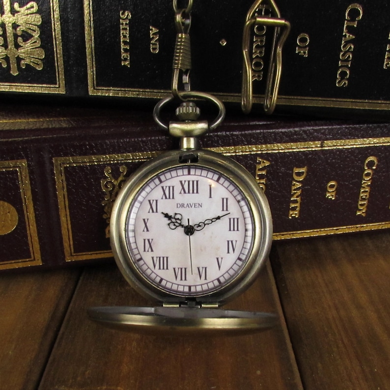 May include: A vintage-style pocket watch with Roman numeral markings on a white face. The watch has a gold-toned finish and is open, revealing the inner workings. The watch is sitting on a stack of books.