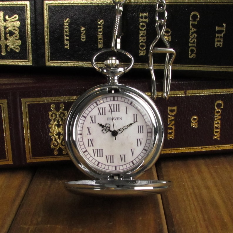 May include: A silver pocket watch with Roman numeral markings on a white face. The watch has a chain attached and is sitting on top of a stack of books. The books have gold lettering on their spines that reads "The Classics of Horror, Stoker and Shelley, Comedy of Dante, Draven".