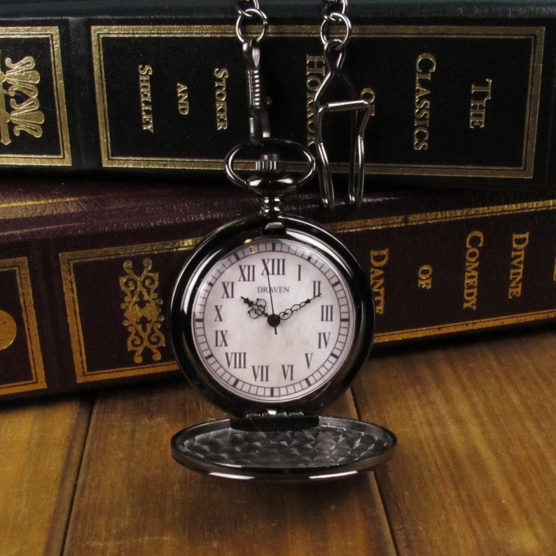 May include: A black pocket watch with Roman numeral markings on a white face. The watch has a chain attached to it and is sitting on top of two books. The books are titled "The Classics" and "Divine Comedy of Dante".