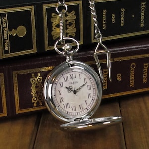 May include: A silver pocket watch with Roman numeral markings on the face. The watch is open and the chain is visible. The watch is sitting on top of a stack of books.