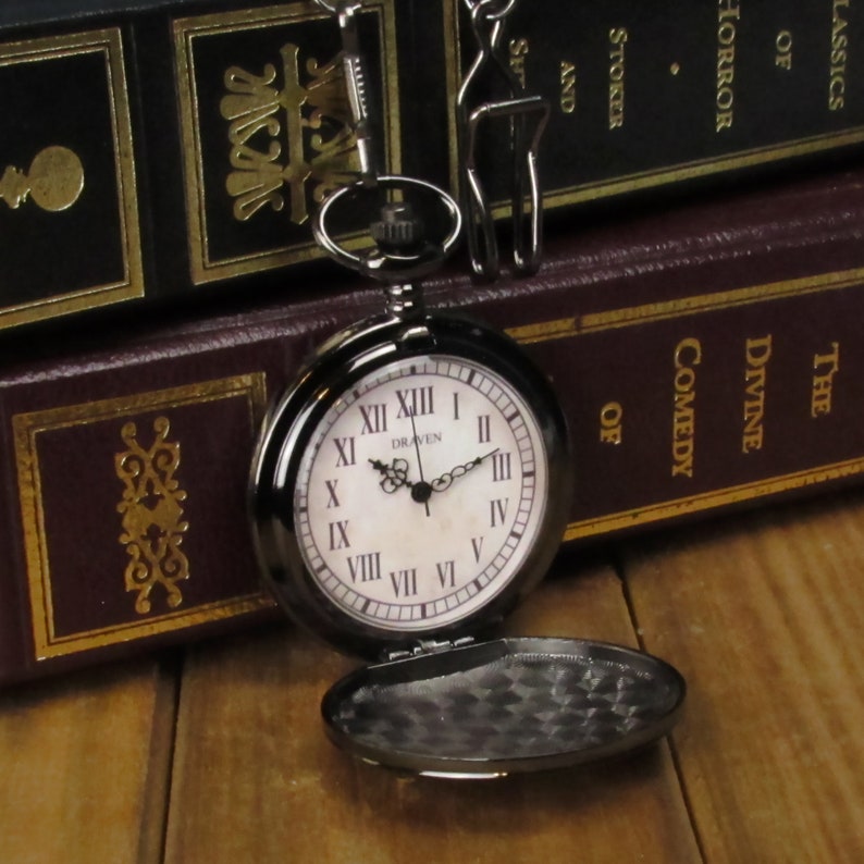 May include: A black pocket watch with Roman numeral markings and a white face. The watch is open and the back is visible. The watch is sitting on top of two books.