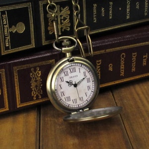 May include: A gold-toned pocket watch with Roman numeral markings on the face. The watch is open and the face reads "DRAVEN". The watch is sitting on top of a stack of books.