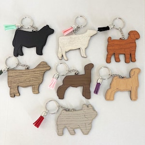 May include: A collection of wooden animal-shaped keychains. Each keychain features a different animal design, including a cow, sheep, and goat. The keychains have silver-colored key rings and colorful tassels in various colors.