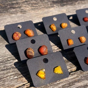 Könnte beinhalten: Ohrstecker aus Bernstein auf schwarzen Karten. Die Bernsteinstücke sind in Gelb-, Orange- und Brauntönen gehalten. Die Ohrringe werden auf einer verwitterten Holzoberfläche präsentiert.