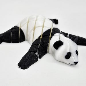 Peut inclure: Figurine de panda imprimée en 3D, en noir et blanc, allongée. Le corps du panda est segmenté, avec des pattes, des oreilles et des marques noires autour des yeux. La figurine est sur une surface blanche.