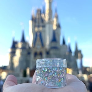 May include: A small, clear plastic jar filled with iridescent glitter, held in an open hand. The glitter includes various shapes and colors, reflecting light. The background features a blurred view of a castle under a blue sky.